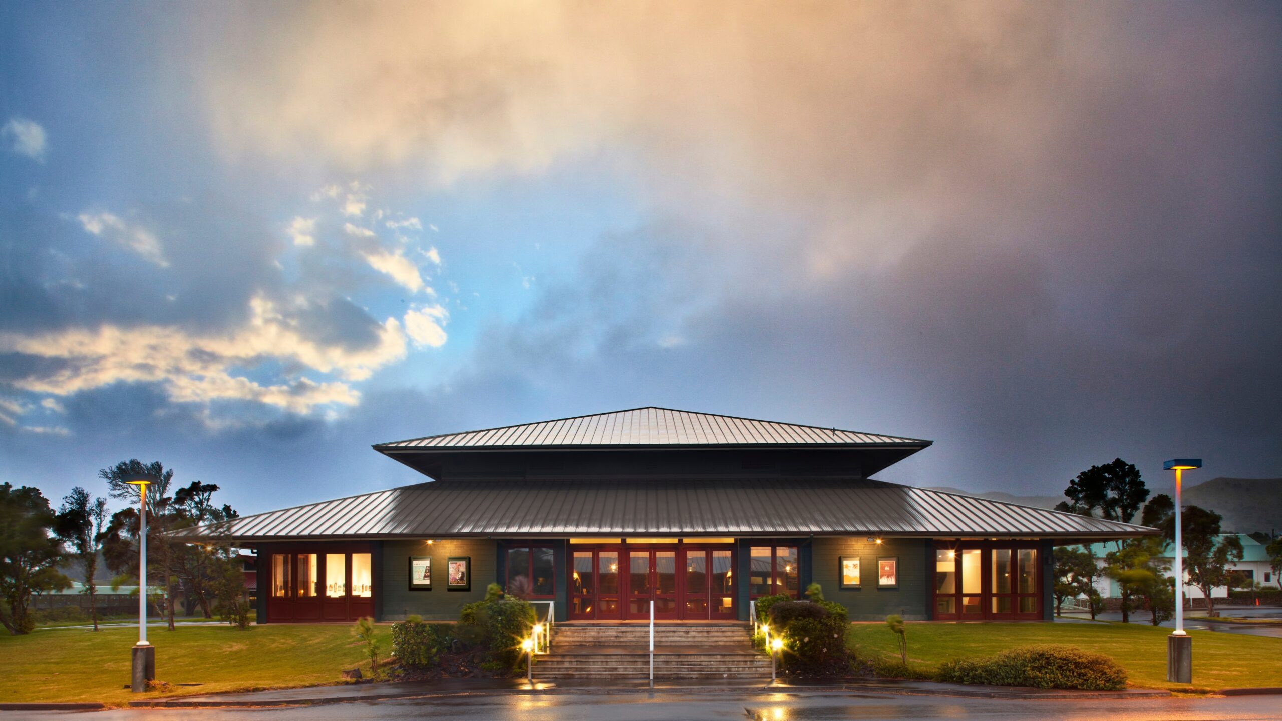 A modern, single-story building with a metal roof, large glass doors, and surrounding greenery under a dramatic cloudy sky at dusk.