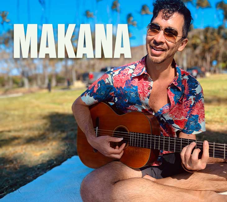 A man wearing sunglasses and a colorful floral shirt plays an acoustic guitar while sitting on a white blanket outside. The word "MAKANA" appears above him.