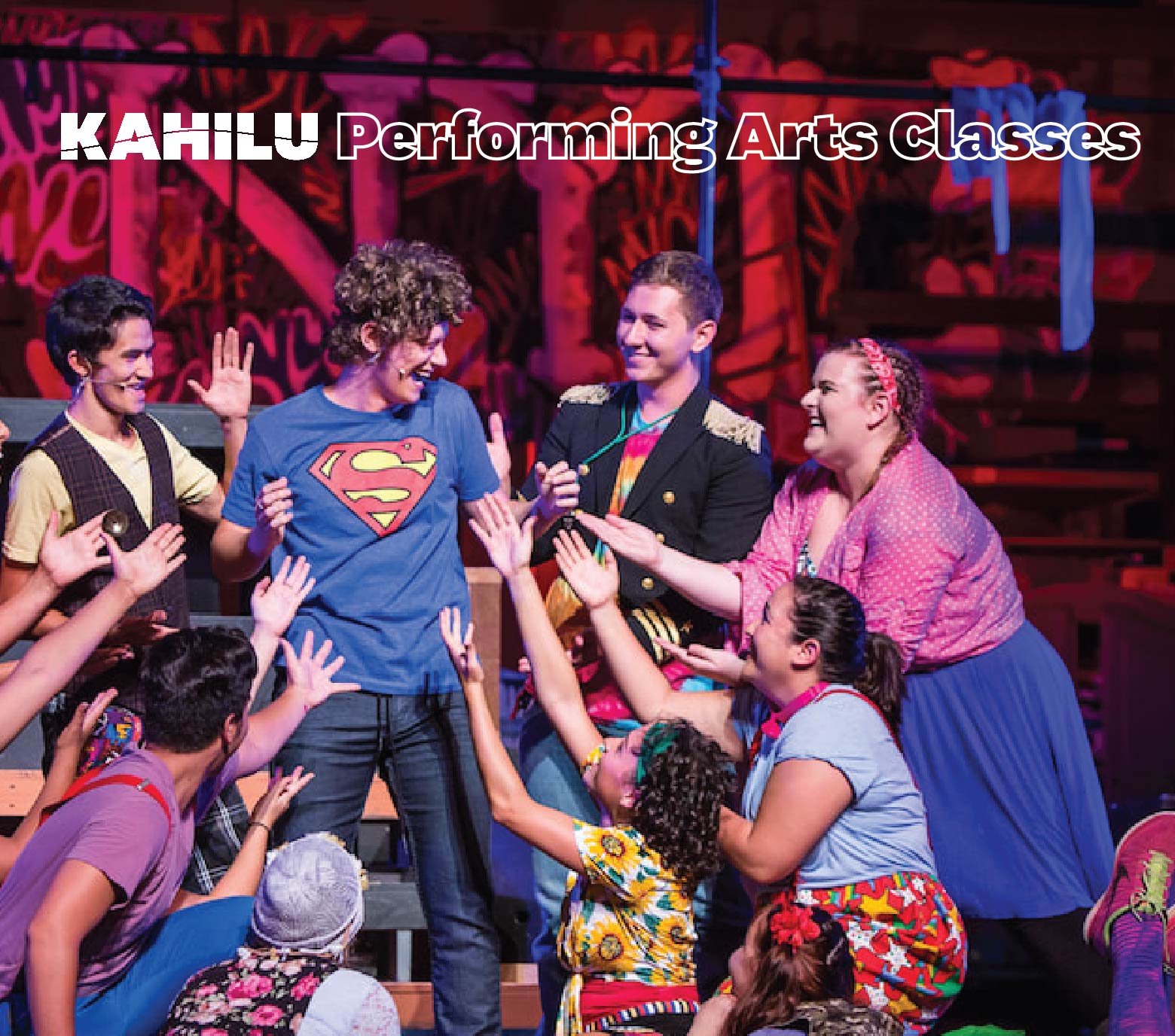 A group of performers in colorful costumes reach towards a person in a Superman t-shirt on stage, with "Kahilu Performing Arts Classes" written at the top.