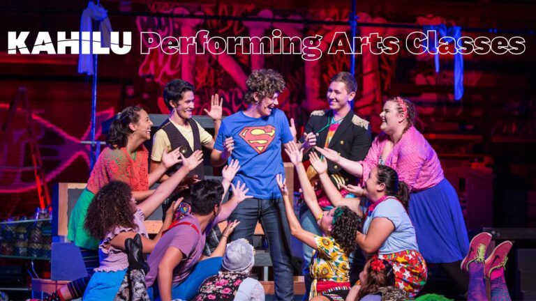 A group of performers on stage enthusiastically gesture towards a person in a Superman shirt, with colorful props and costumes surrounding them. Text reads "Kahilu Performing Arts Classes and Workshops.