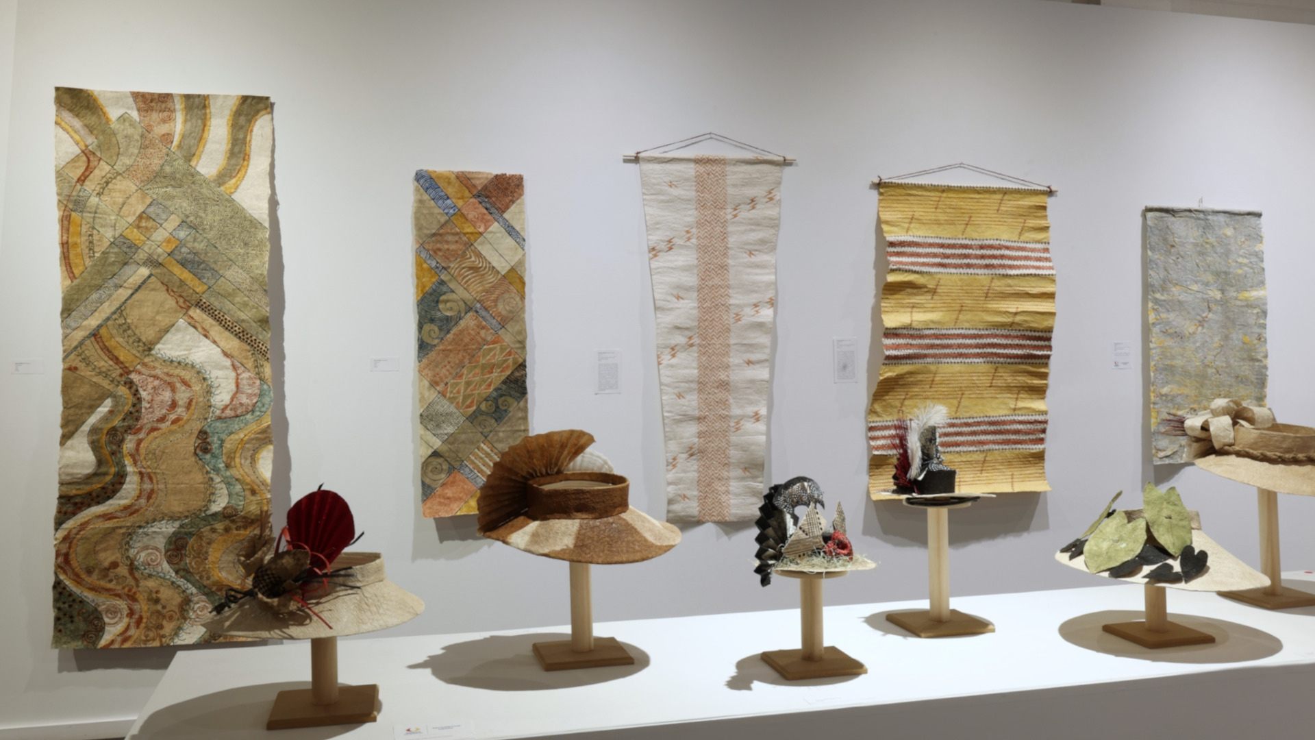 Display of textile art and hats on pedestals in a gallery, featuring various patterns and colors, mounted on a white wall.