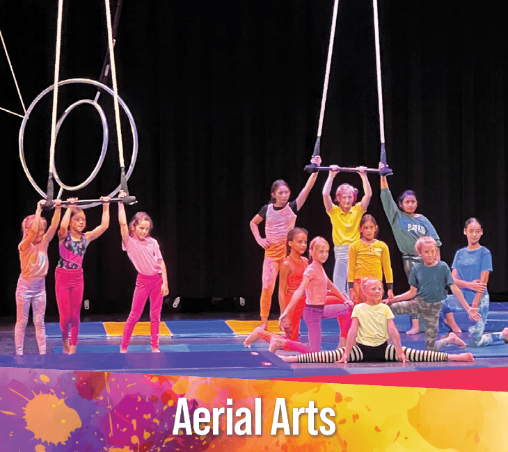A group of children pose on stage with aerial hoops and a trapeze, wearing colorful outfits, beneath a banner reading "Aerial Arts.