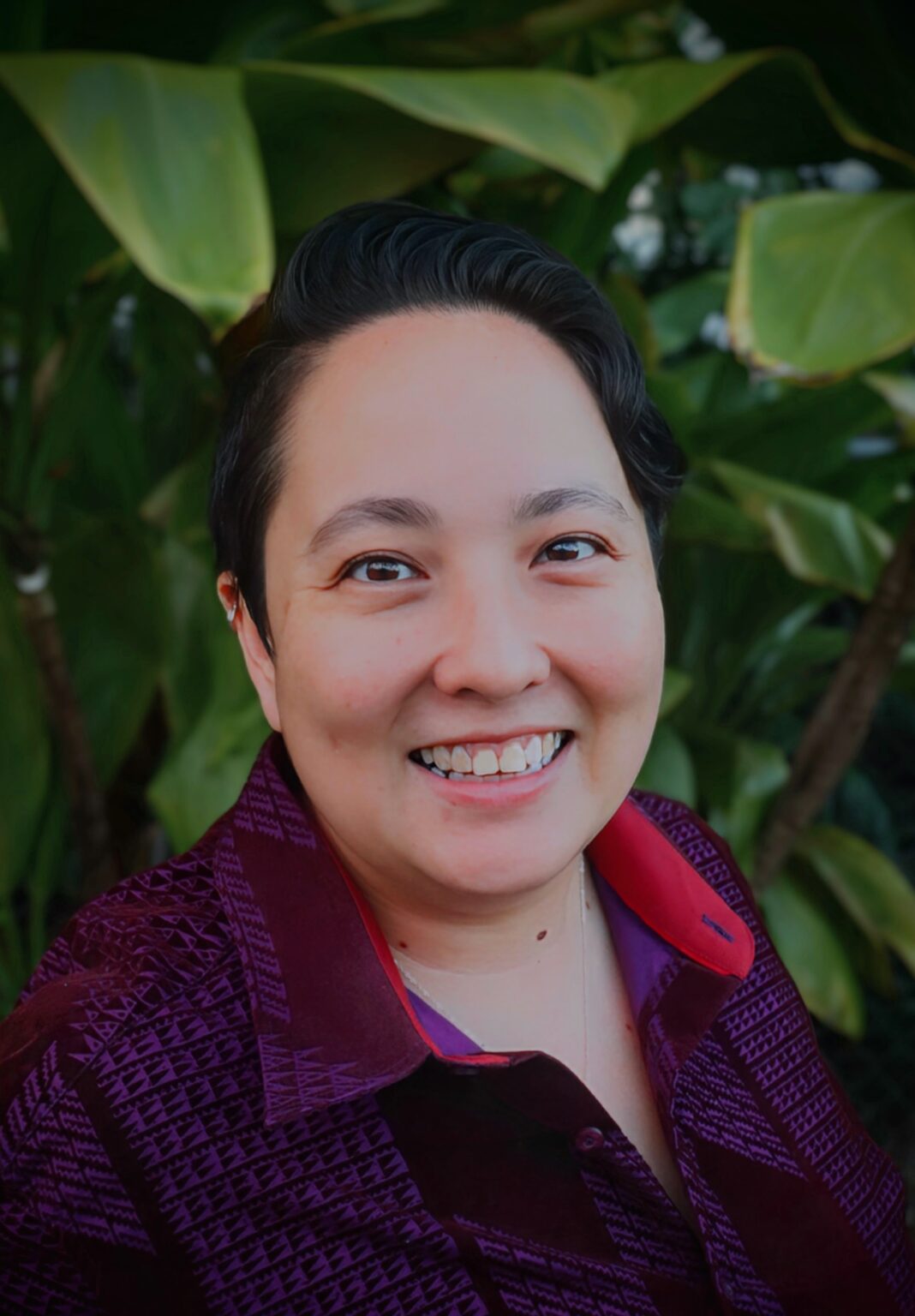 A person with short dark hair smiles at the camera, wearing a maroon patterned shirt, with large green leaves in the background.