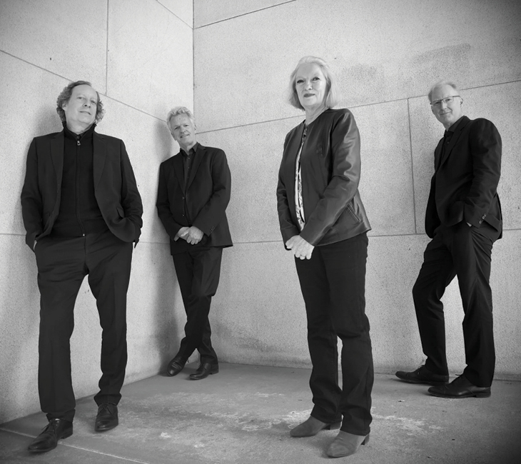 Four adults in dark clothing stand against a light stone wall, posing for a group photo. The image is in black and white.