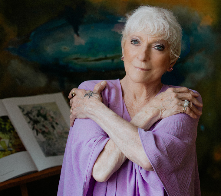 Elderly woman with short white hair wearing a light purple shawl, arms crossed over her chest, standing in front of an abstract painting and an open book.