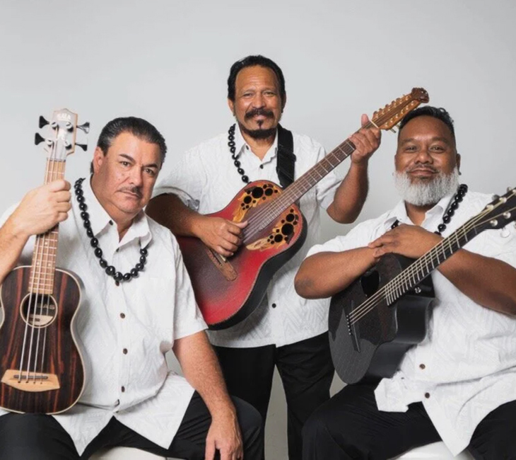 Three men wearing white shirts and black pants pose with stringed instruments against a plain background.