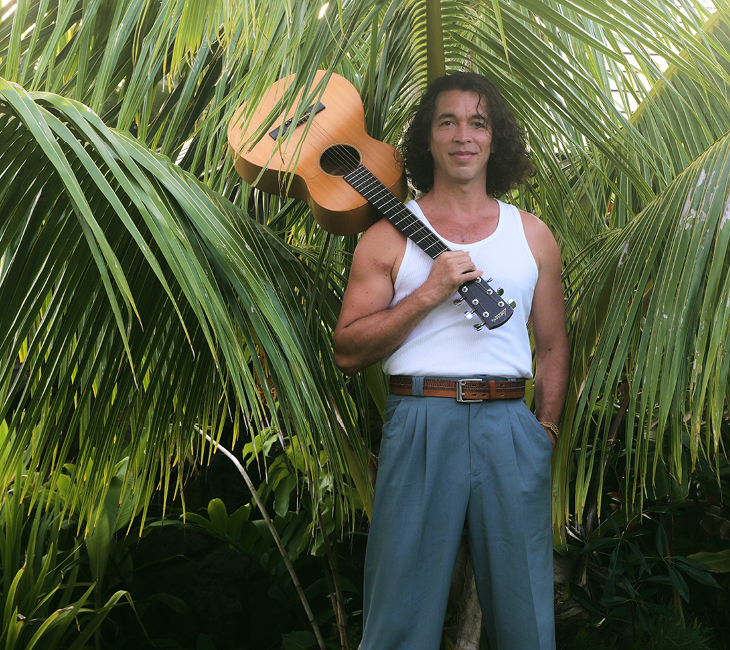 A man in a white tank top and blue pants stands among palm leaves, holding an acoustic guitar over his shoulder.