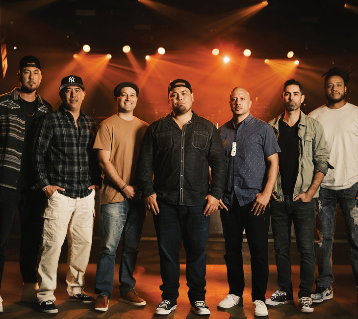 Seven men stand side by side on stage with orange lights in the background, all facing the camera and dressed in casual clothing.
