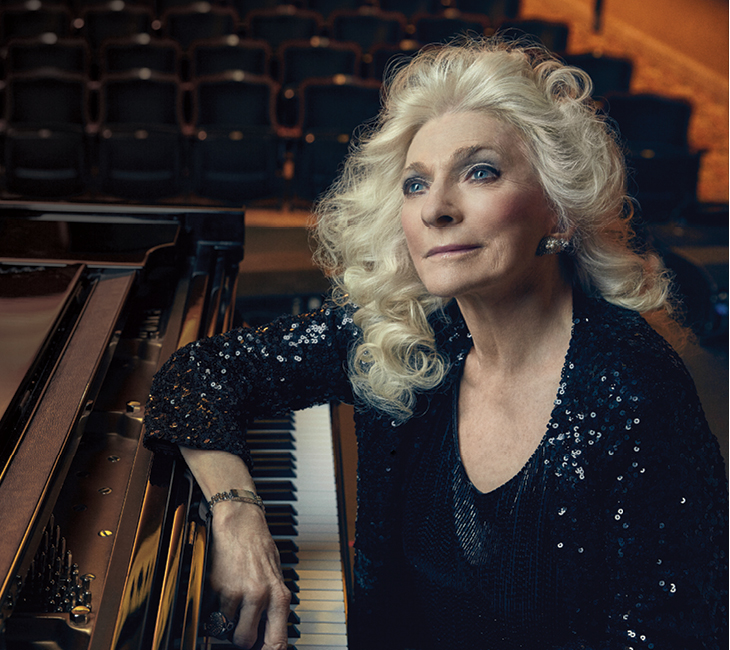 An older woman with long white hair and a sequined black outfit sits at a grand piano on a stage with empty seats in the background.