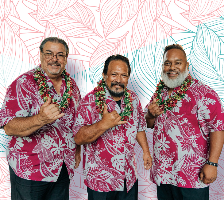 Three men wearing matching red Hawaiian shirts and floral leis stand in front of a white and pink leaf-patterned backdrop, smiling and making a shaka hand gesture.