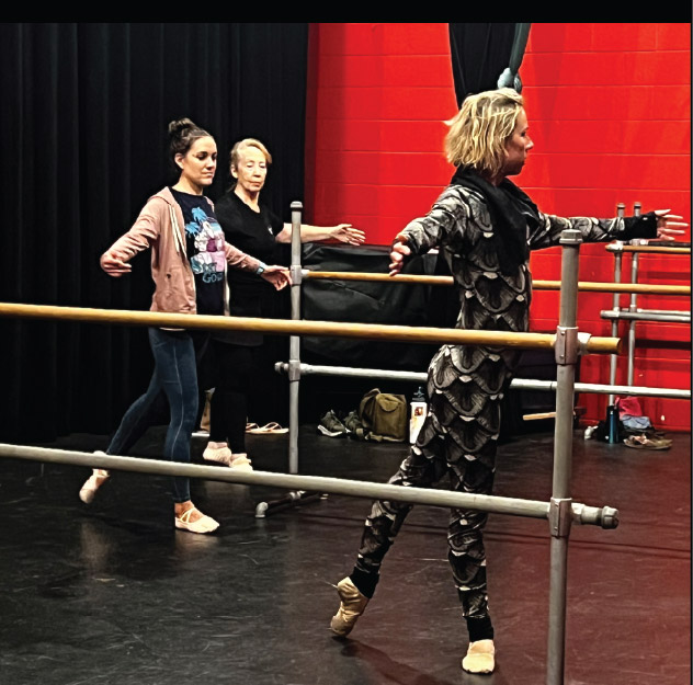 Three women practice ballet at a barre in a dance studio with red and black walls. Two are in casual wear, one is in a patterned leotard and pointe shoes, demonstrating a ballet pose.