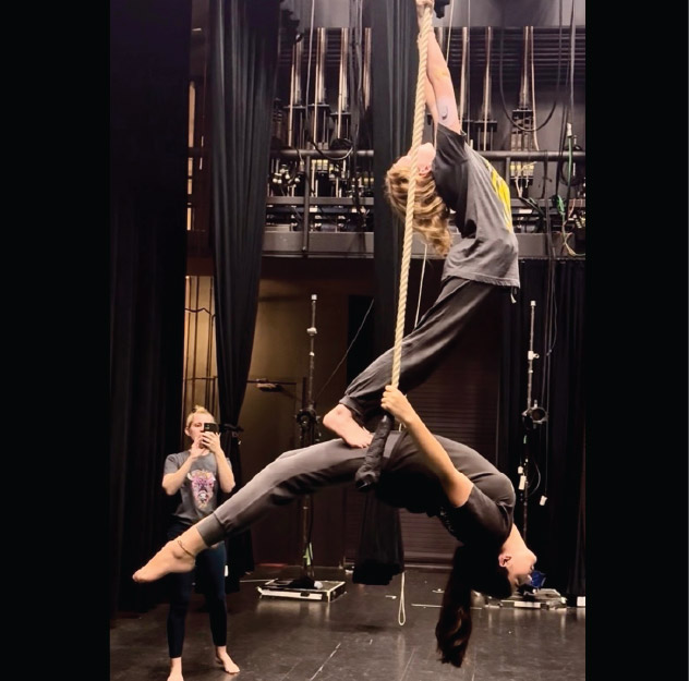 Two people perform an aerial routine on a rope while a third person stands nearby taking a photo on a stage with theater rigging visible in the background.