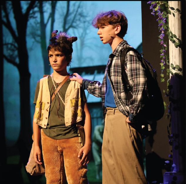 Two boys in costume perform on stage; one wears animal ears and fur pants, the other wears a plaid shirt and khakis, with a forest backdrop behind them.
