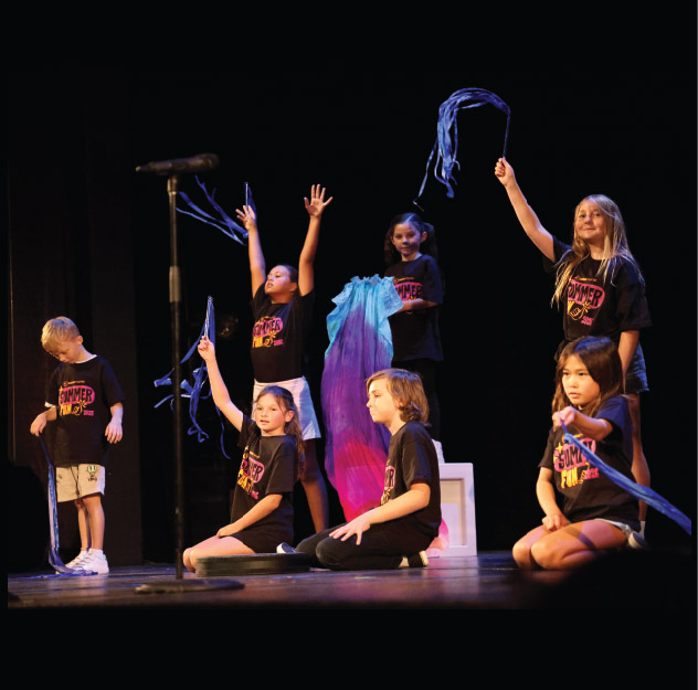 Seven children wearing black "Summer Stage" shirts perform onstage, holding blue ribbons and fabric props, with a microphone stand visible on the left.