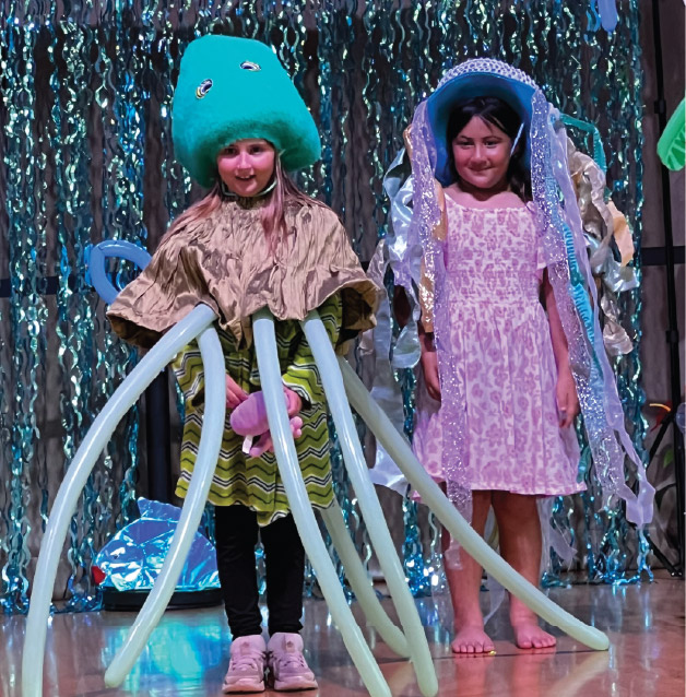 Two children in sea creature costumes stand on stage; one is dressed as an octopus with balloon arms, the other as a jellyfish with a shiny headpiece and streamers, against a decorated backdrop.