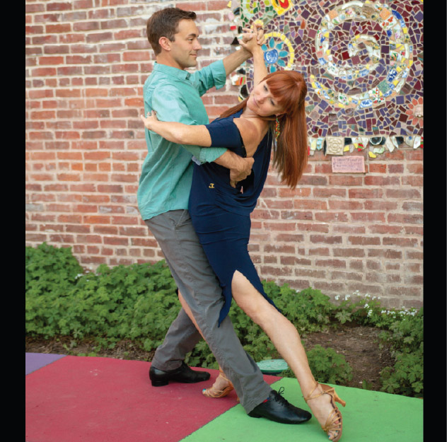 A man and woman dance together outdoors, performing a dip on a colorful mat near a brick wall with a mosaic artwork.
