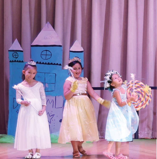 Three children in princess costumes stand on stage in front of a blue castle backdrop, smiling and holding props.