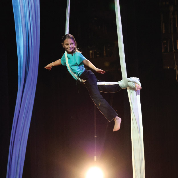 A young person performs an aerial silk routine, hanging by one leg and one arm, with stage lights shining from below against a dark background.