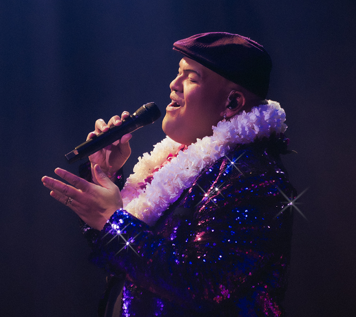 A person wearing a dark cap, sparkly purple jacket, and white flower lei sings into a microphone on stage under soft lighting.