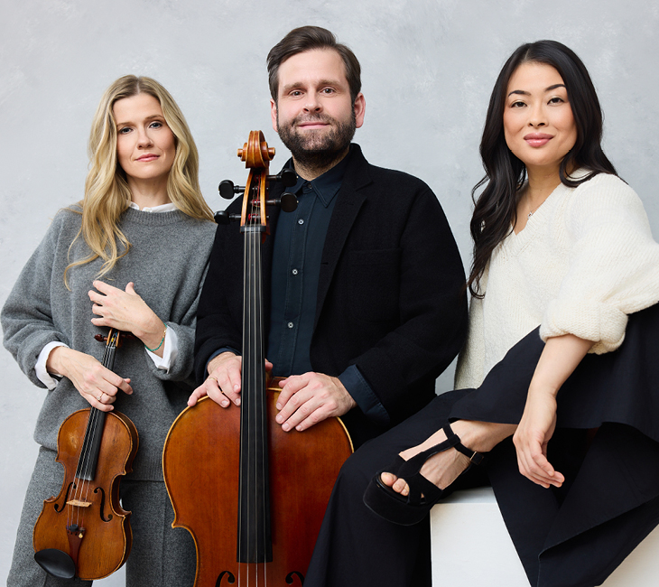 Three musicians pose together; the woman on the left holds a violin, the man in the center sits with a cello, and the woman on the right sits with one leg crossed, all facing the camera.