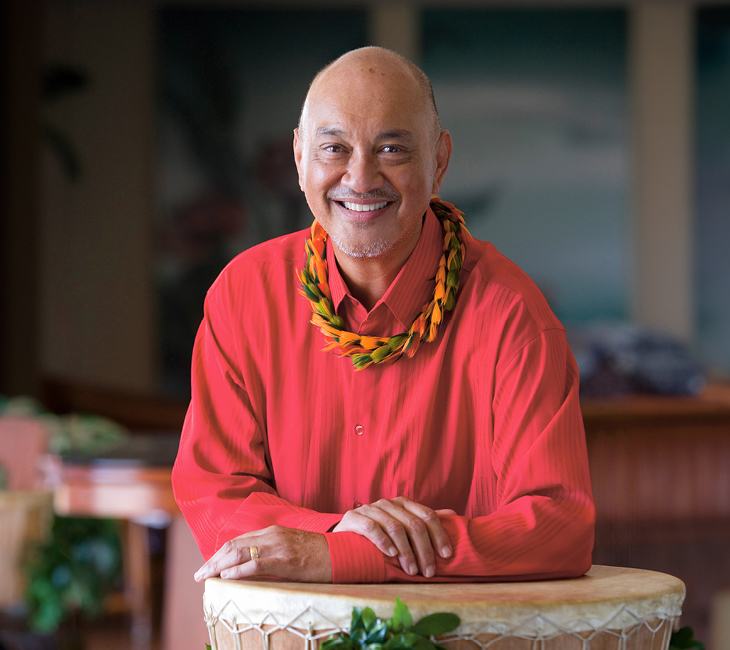 A man in a red shirt and yellow lei smiles, leaning on a drum in an indoor setting with plants and blurred background.