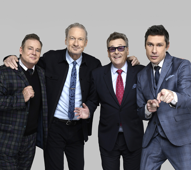 Four men in suits stand together against a plain gray background, smiling and posing, with one man pointing toward the camera.