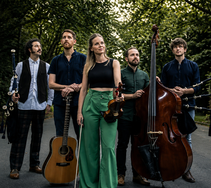 Five musicians stand outdoors with their instruments: bagpipes, acoustic guitar, violin, upright bass, and mandolin, surrounded by trees.