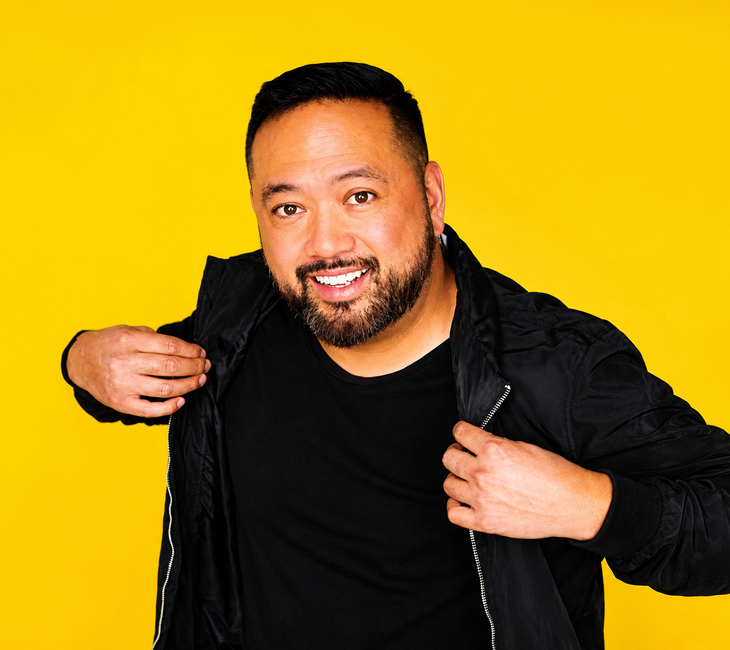 A man with a beard smiles at the camera while adjusting his black jacket. He stands in front of a solid bright yellow background.