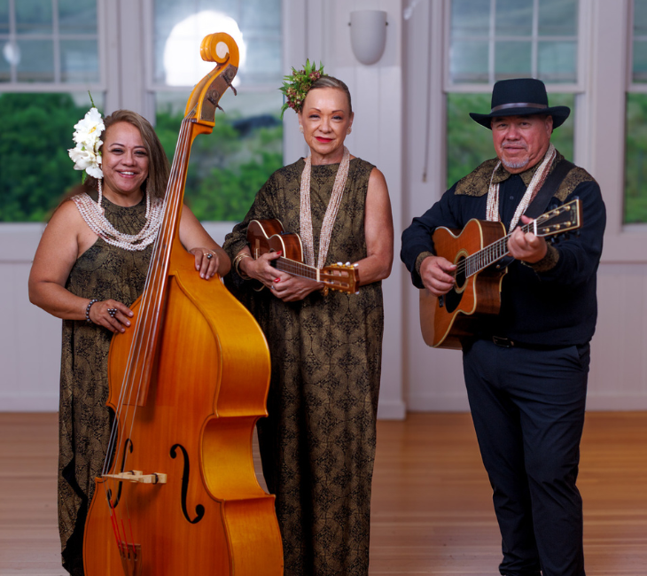 Three musicians in patterned outfits stand indoors with a double bass, a ukulele, and a guitar, smiling for the camera.