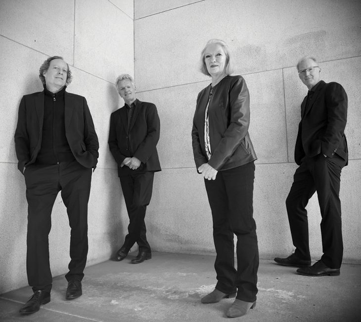 Four adults in dark clothing stand against a light stone wall, posing for a group photo. The image is in black and white.