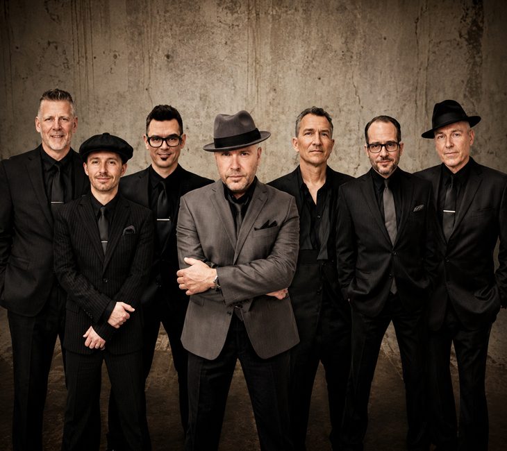 Seven men wearing dark suits and hats stand in a row against a textured gray wall, facing the camera with neutral expressions.