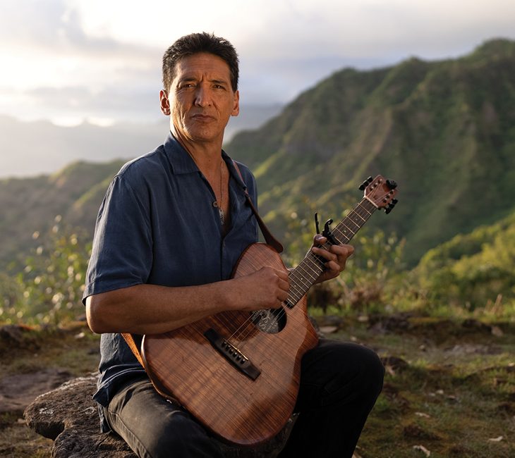 A man sits on a rock outdoors, playing an acoustic guitar, with green hills and cloudy sky in the background.