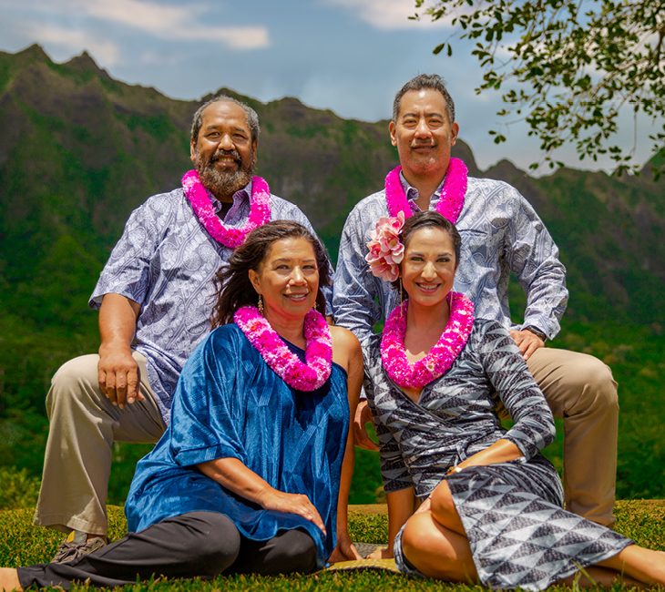 Four adults wearing leis pose outdoors in front of green mountains, two seated and two standing behind them, smiling at the camera.