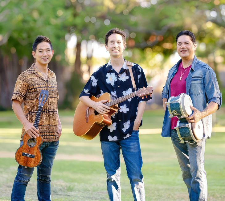 Three men standing outdoors, each holding a musical instrument: a ukulele, a guitar, and a set of bongos. They are smiling and dressed in casual, colorful shirts and jeans.