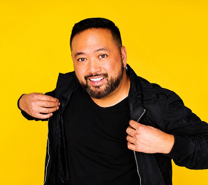 A man with a beard smiles at the camera while adjusting his black jacket. He stands in front of a solid bright yellow background.