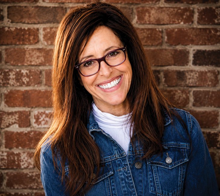 Woman with long brown hair and glasses smiles at the camera, wearing a denim jacket and white shirt, standing in front of a brick wall.