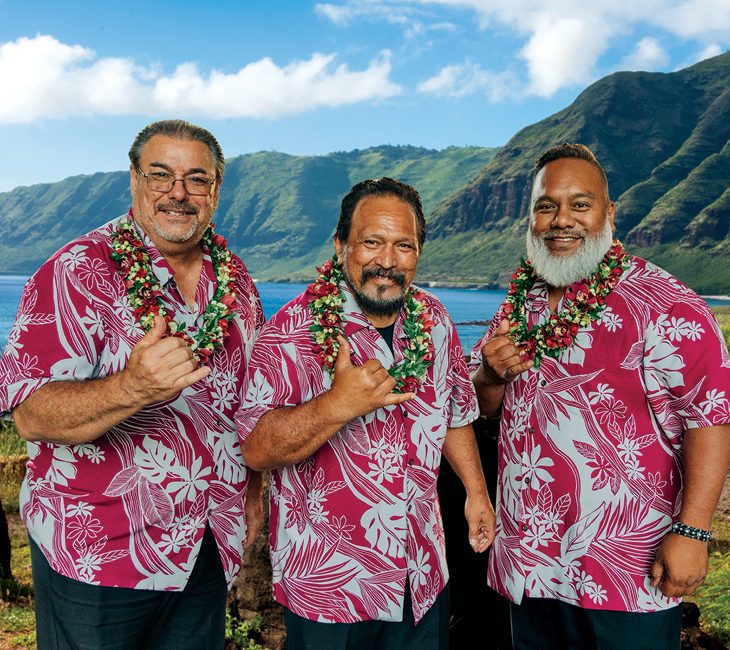 Three men wearing matching red Hawaiian shirts and flower leis make shaka hand signs, standing outdoors with mountains and ocean in the background.