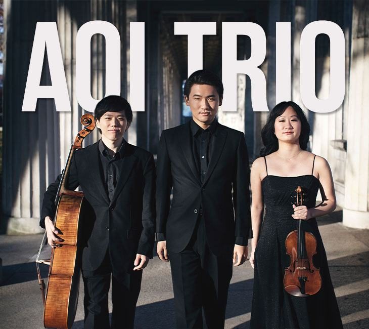 Three musicians dressed in black formal attire stand in front of large letters spelling "AOI TRIO." One holds a cello, one is empty-handed, and the third holds a violin.