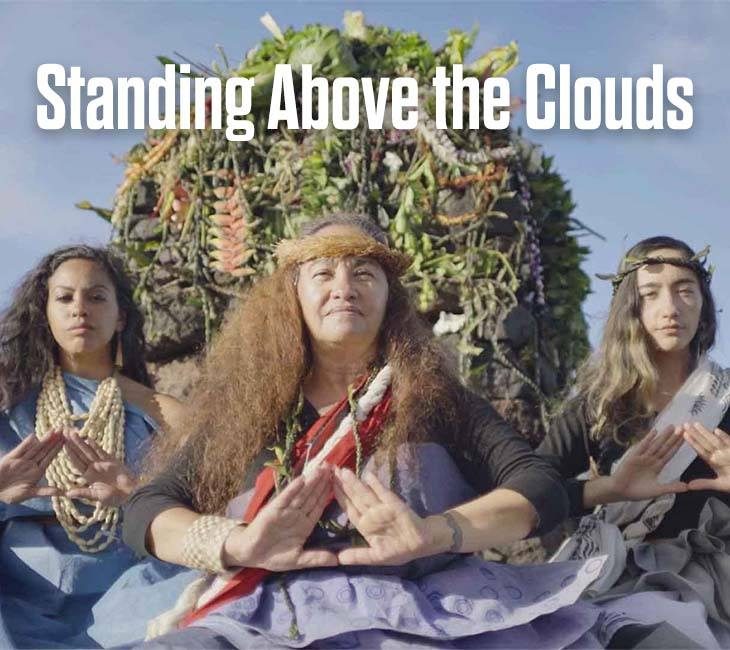 Three people wearing traditional attire and accessories are seated in front of a lush backdrop. They are holding their hands in symbolic positions. Text above reads, "Standing Above the Clouds.