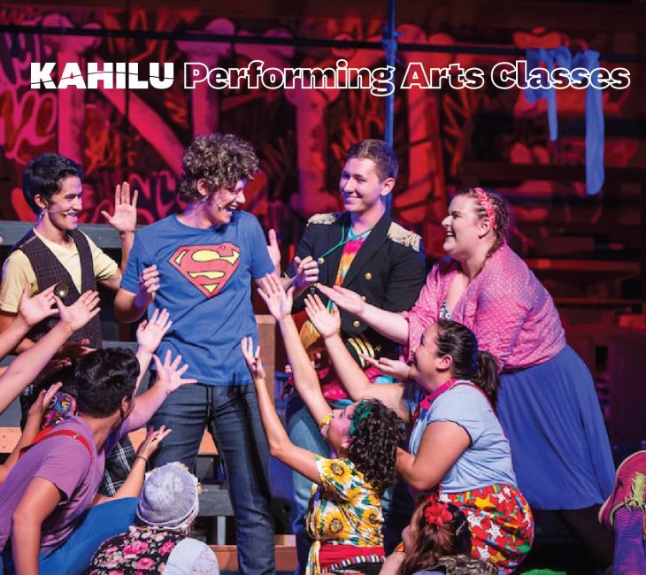 A group of performers in colorful costumes reach towards a person in a Superman t-shirt on stage, with "Kahilu Performing Arts Classes" written at the top.