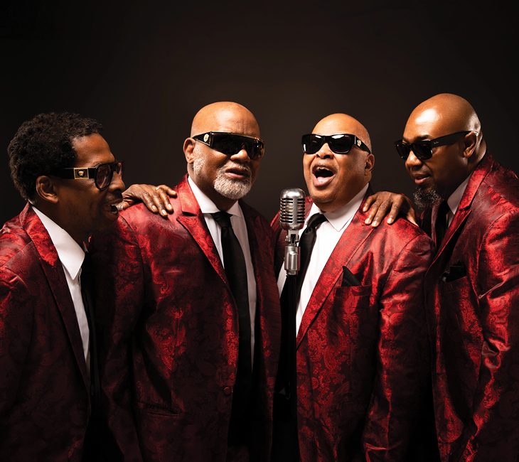 Four men in matching red patterned suits and sunglasses sing together around a vintage microphone against a dark background.