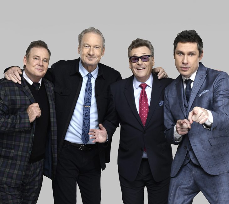 Four men in suits stand together against a plain gray background, smiling and posing, with one man pointing toward the camera.
